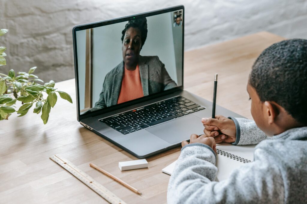 Back view of crop anonymous smart diligent African American boy writing in notebook while teacher dictating online at laptop